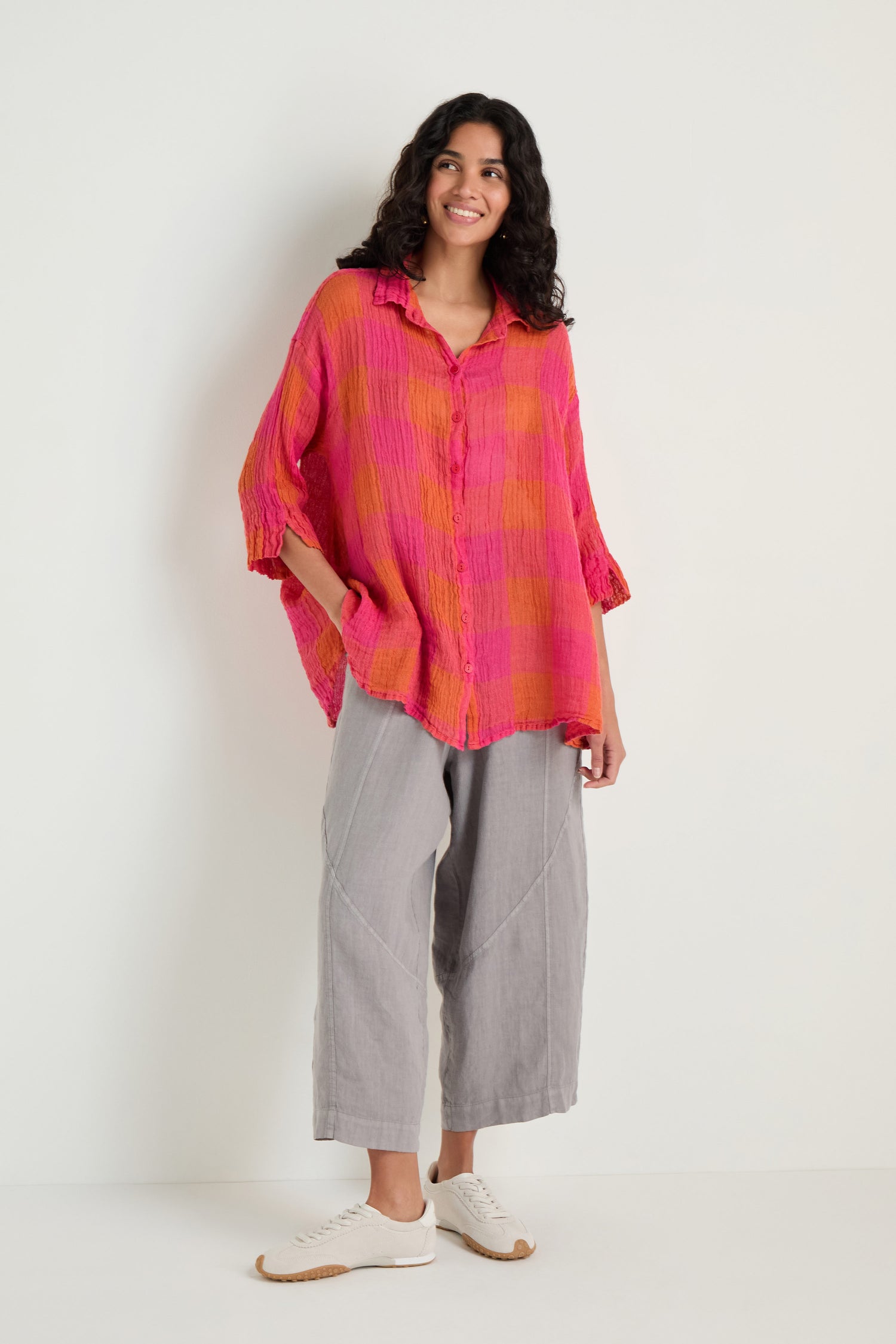 Person stands against a plain background, wearing the Large Check Linen Swing Shirt in pink and orange, paired with light grey wide-leg pants and white sneakers, hands in pockets, sporting a relaxed expression.