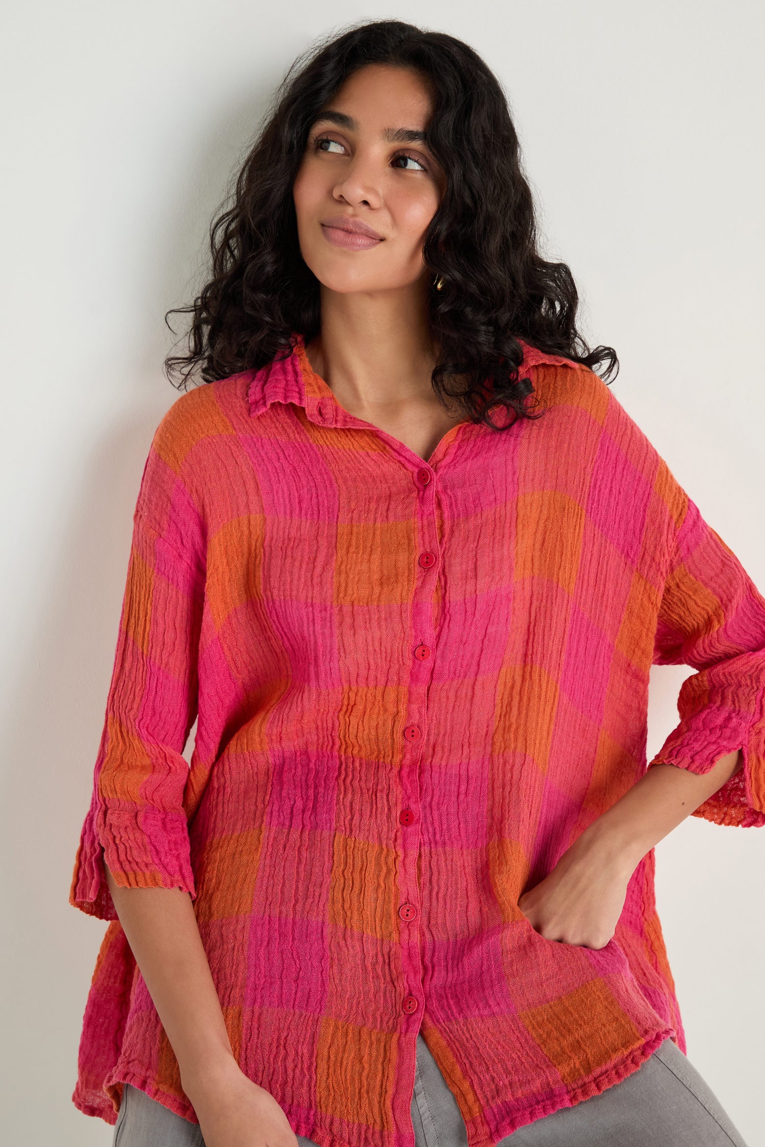 A woman with wavy hair stands against a plain light wall, wearing the Large Check Linen Swing Shirt in a relaxed fit, featuring pink and orange checks.