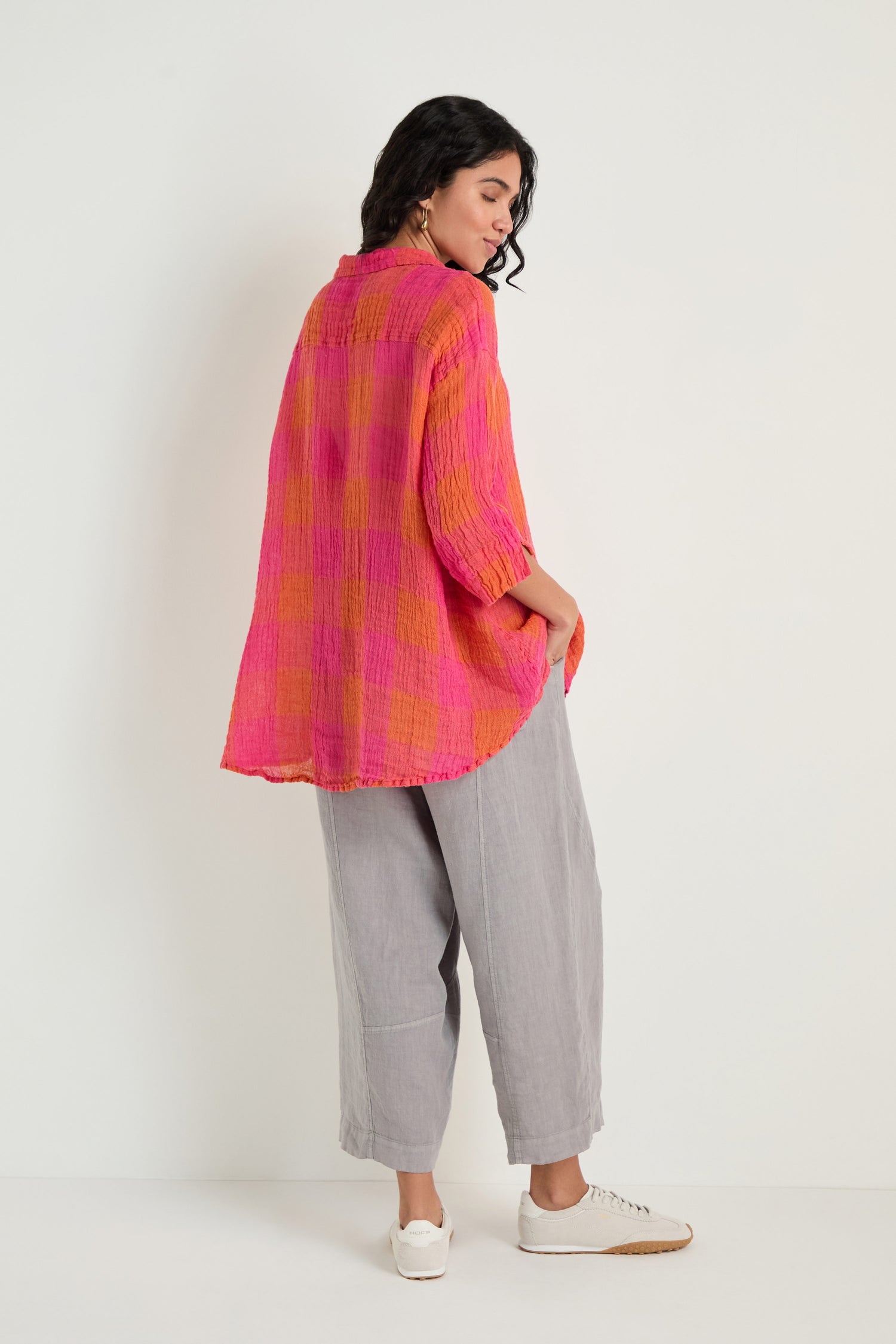A person stands against a white wall, wearing the Large Check Linen Swing Shirt with a pink and orange check pattern, paired with light gray wide-leg pants and white sneakers.