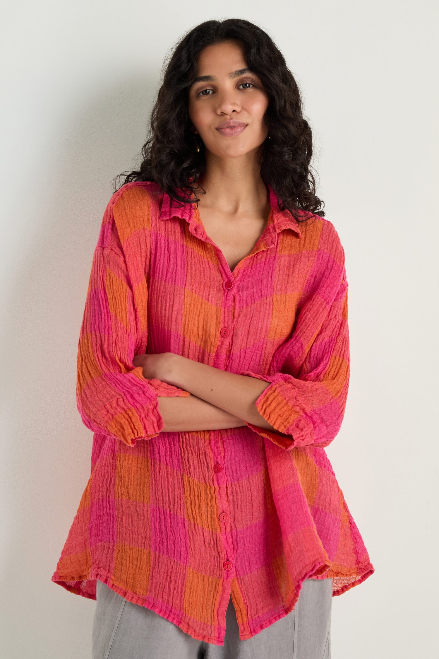 A woman with long curly hair wears the Large Check Linen Swing Shirt in pink and orange, paired with light gray pants, standing against a plain light background.