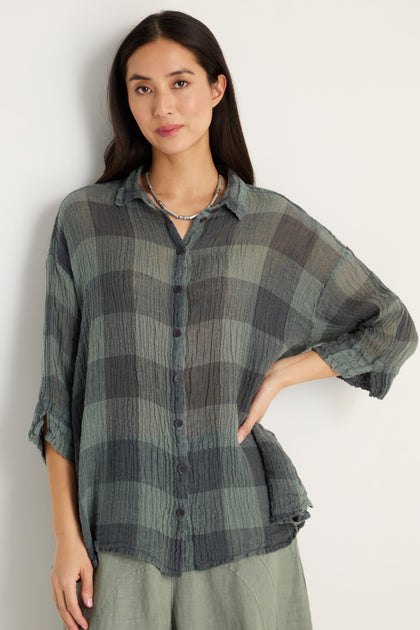 A woman wears the Large Check Sage Linen Swing Shirt—a breathable, lightweight, long-sleeve plaid top with a relaxed swing fit in green tones—paired with light pants, standing against a plain white background.