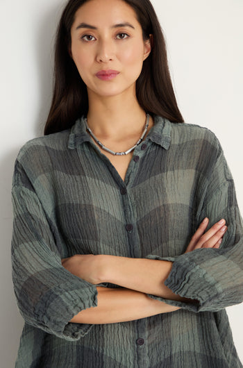 A woman with long dark hair stands with arms crossed, wearing the Large Check Sage Linen Swing Shirt and a necklace, facing the camera against a plain light background.