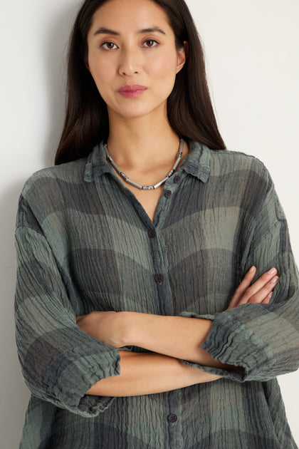 A woman with long dark hair stands with arms crossed, wearing the Large Check Sage Linen Swing Shirt and a necklace, facing the camera against a plain light background.