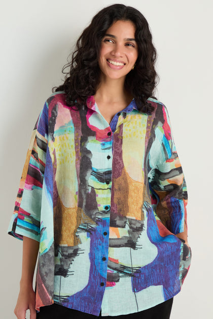 Smiling, a woman with long dark hair wears the Abstract Print Flared Shirt—a loose, breathable linen piece featuring an abstract design—while standing against a plain light wall.