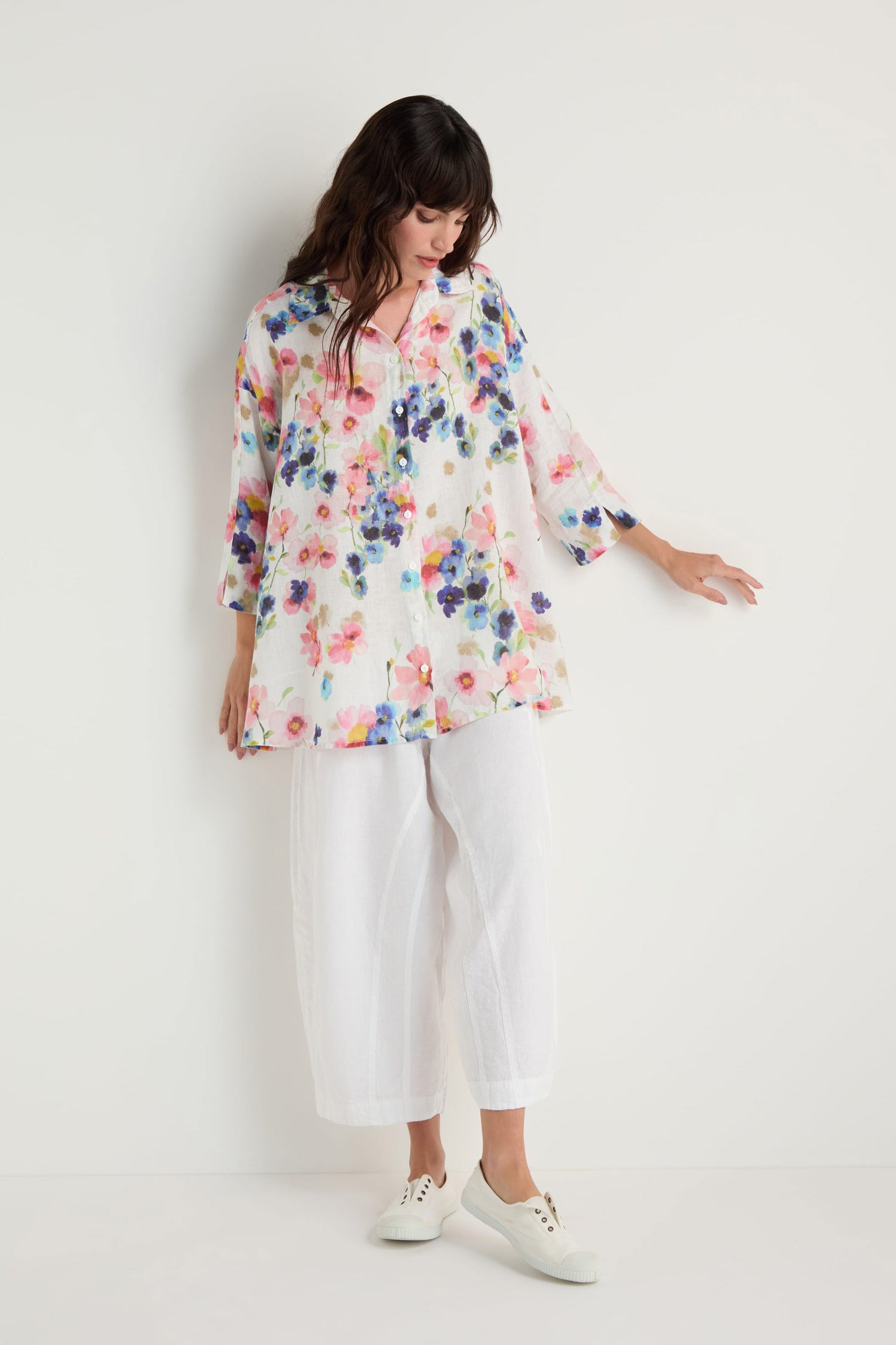 Woman stands against a white wall, wearing the Primrose Print Flared Linen Shirt, white wide-leg pants, and white sneakers. She looks down and touches the wall with one hand.