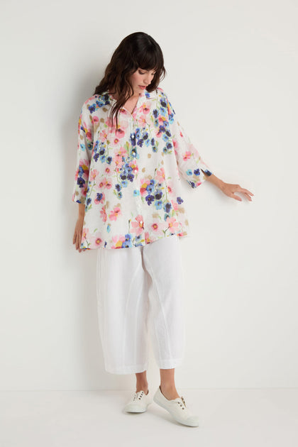 Woman stands against a white wall, wearing the Primrose Print Flared Linen Shirt, white wide-leg pants, and white sneakers. She looks down and touches the wall with one hand.