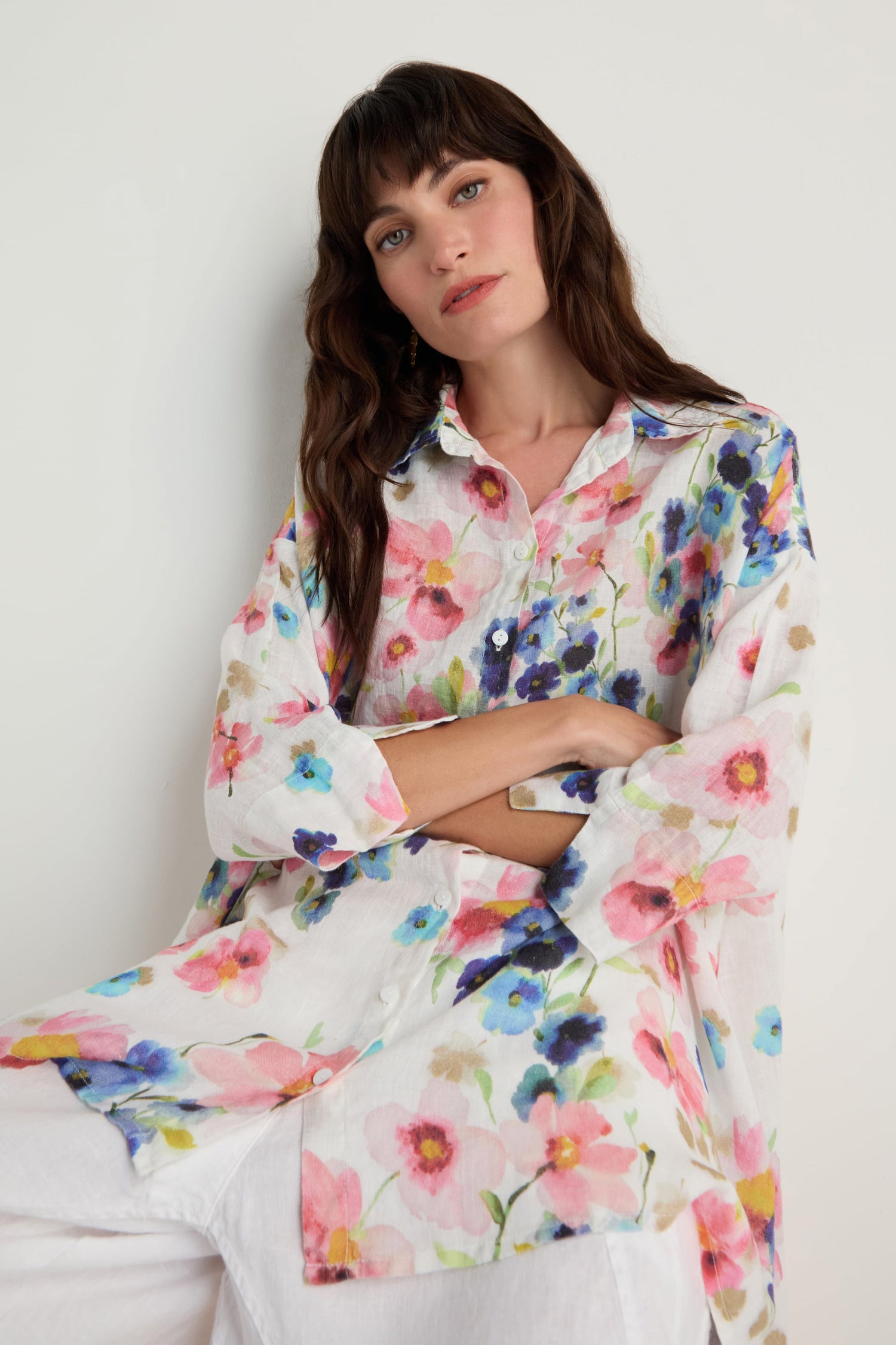 Woman wearing the Primrose Print Flared Linen Shirt, a relaxed fit, long-sleeved white linen shirt with a colorful floral print, sits against a plain light background with arms crossed.