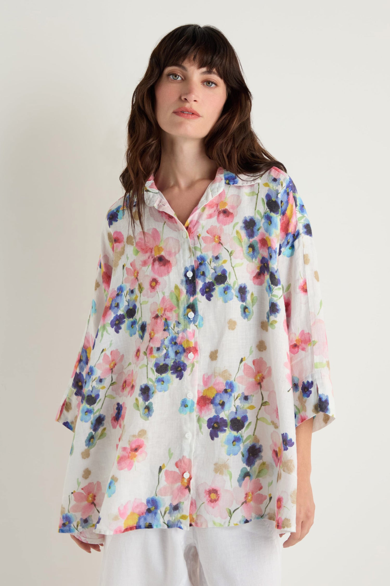 A woman models the Primrose Print Flared Linen Shirt, a relaxed-fit white linen top with a vibrant floral pattern, standing against a plain light background.