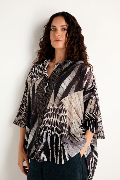 A woman with long curly hair stands against a plain light-colored wall, wearing the Patchwork Print Flared Shirt and dark pants, looking at the camera with a neutral expression.