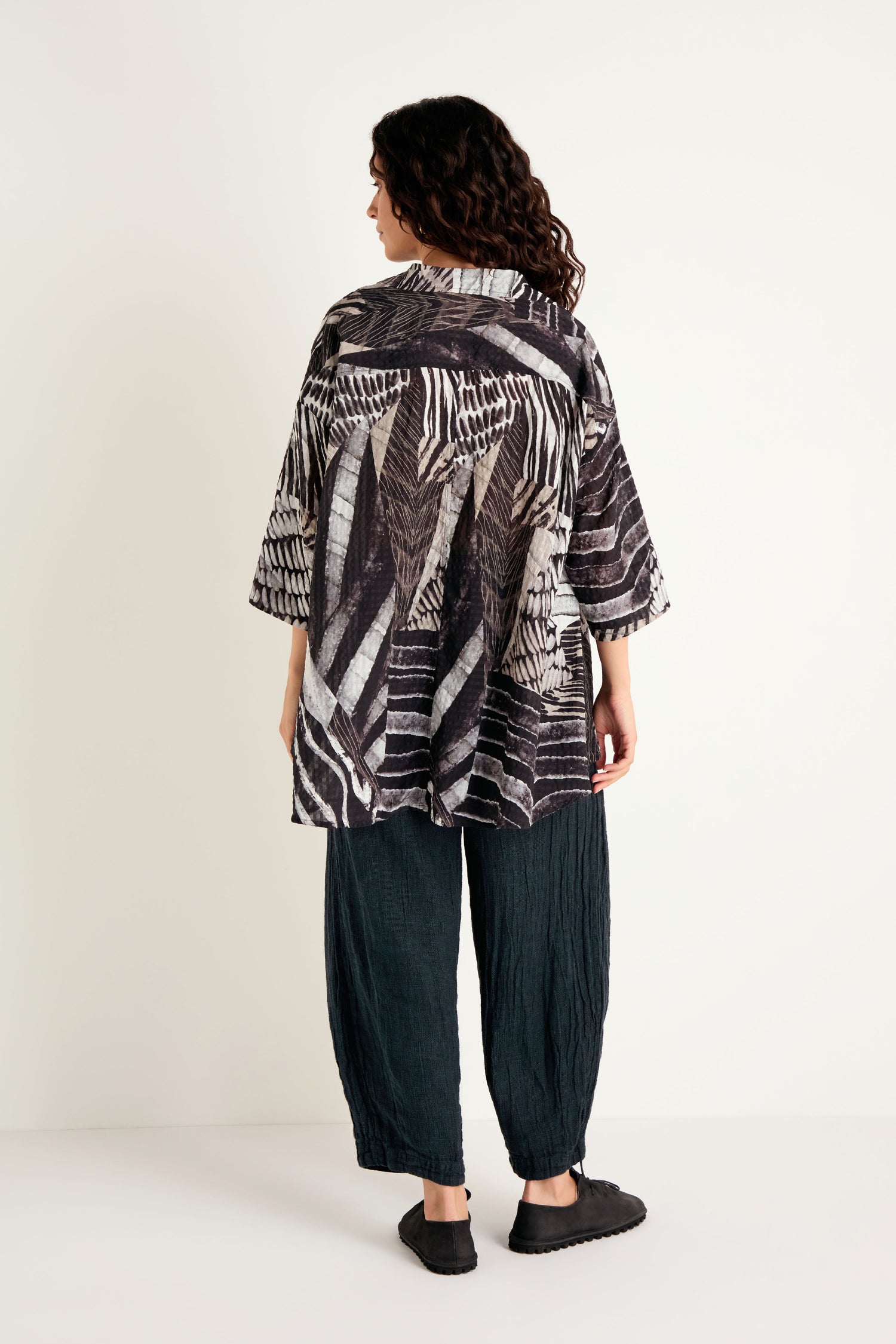 A person stands with their back to the camera, wearing the Patchwork Print Flared Shirt in a black and white silk cotton blend, styled with loose dark pants and black shoes against a plain white background.