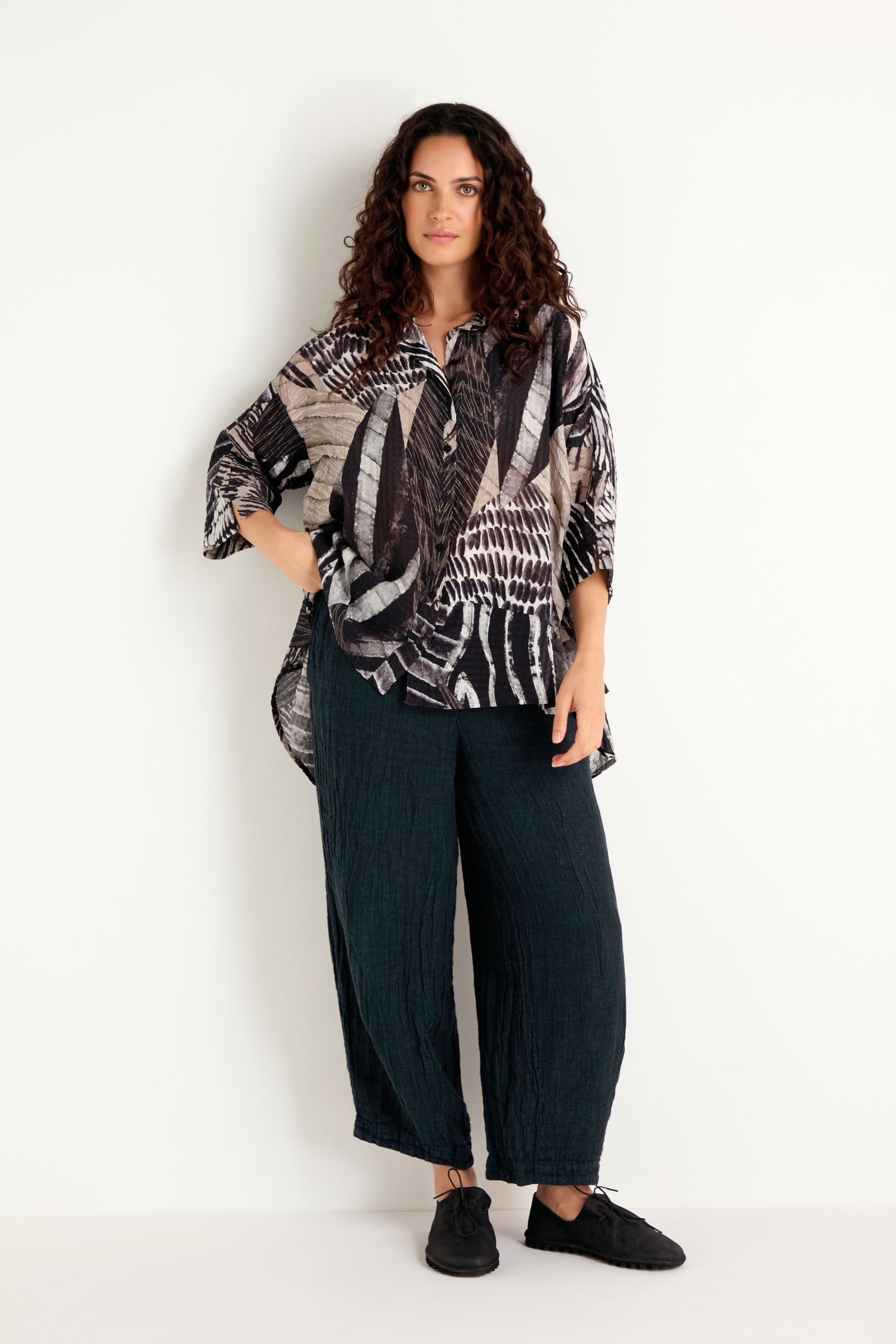 A woman with curly hair stands against a white wall, wearing the Patchwork Print Flared Shirt in a loose-fitting flared silhouette, paired with dark wide-leg pants and black shoes.
