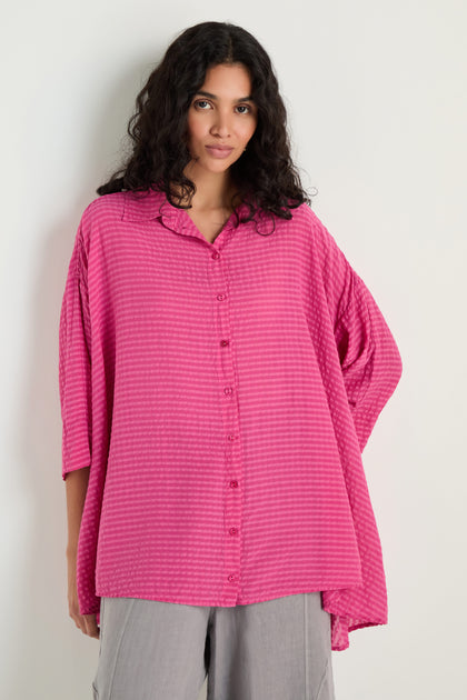 A woman wears the Seersucker Silk Blend Flared Shirt in fuchsia with subtle stripes and long sleeves, paired with light gray pants, standing against a plain light background.