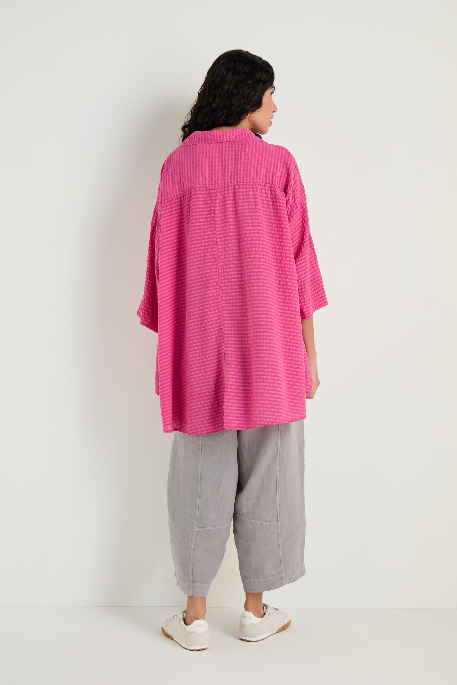 A person faces away against a white wall, wearing the Seersucker Silk Blend Flared Shirt in fuchsia, paired with gray wide-leg pants and white shoes.