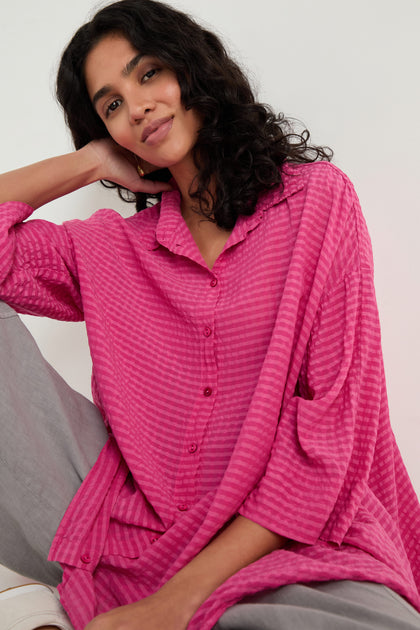 A person with dark curly hair sits casually against a plain background, wearing the Seersucker Silk Blend Flared Shirt in fuchsia and light gray pants.