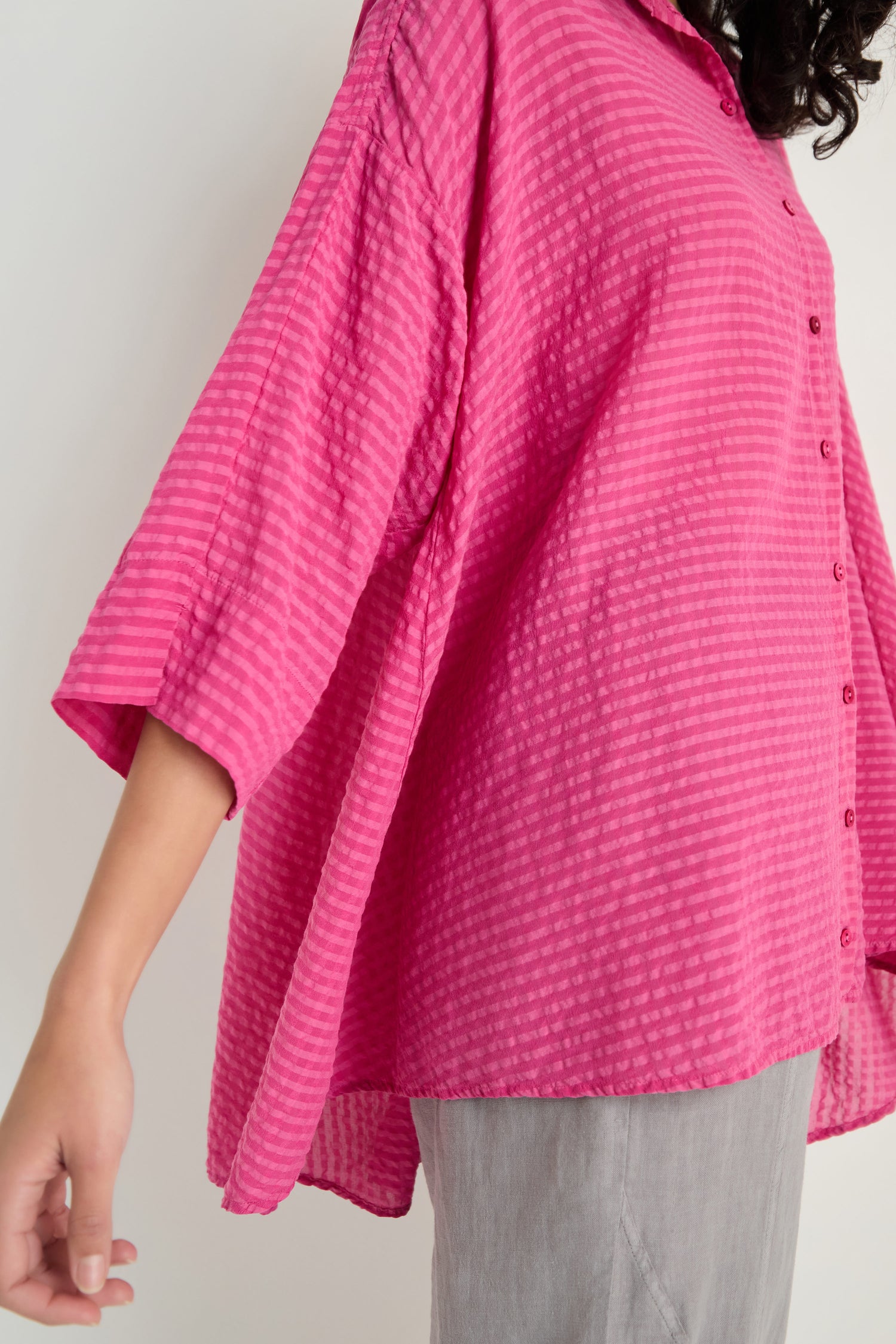 Model wears the Seersucker Silk Blend Flared Shirt in fuchsia, featuring a loose fit, wide sleeves, and textured finish, paired with light gray pants. Shown from the side.