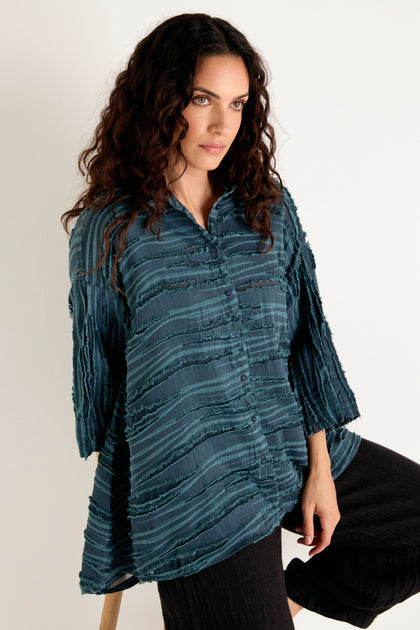 Woman with long curly hair wearing the Deep Turquoise Textured Stripe Flared Shirt—a linen silk cotton blend in a textured, button-up tunic style—paired with black pants, standing against a plain light background.