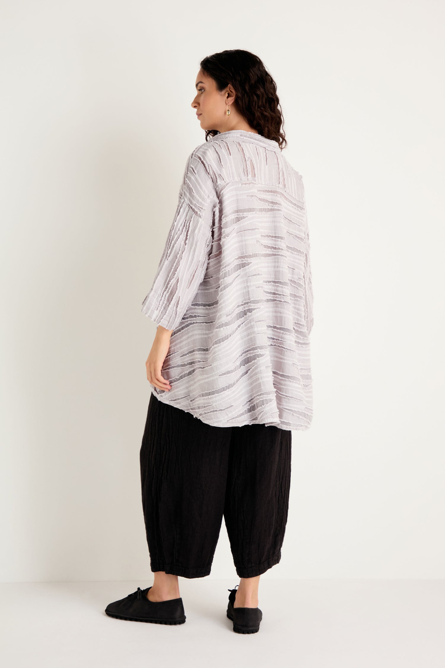 Person standing against a white background, wearing the Pearl Textured Stripe Flared Shirt—a loose, light-colored top with three-quarter sleeves—and black wide-leg pants, facing slightly away from the camera.