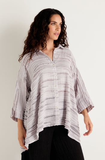 A woman with long curly hair wears the Pearl Textured Stripe Flared Shirt, a loose, light-colored top crafted from a linen silk cotton blend with an asymmetrical hem and textured detail, paired with black pants as she stands against a plain light background.