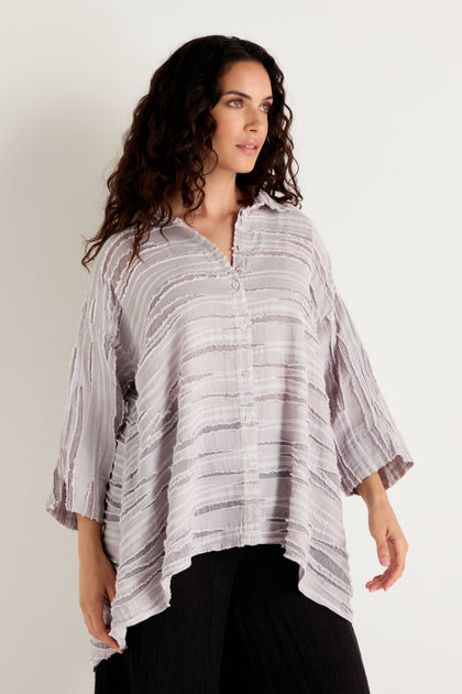 A woman with long curly hair wears the Pearl Textured Stripe Flared Shirt, a loose, light-colored top crafted from a linen silk cotton blend with an asymmetrical hem and textured detail, paired with black pants as she stands against a plain light background.
