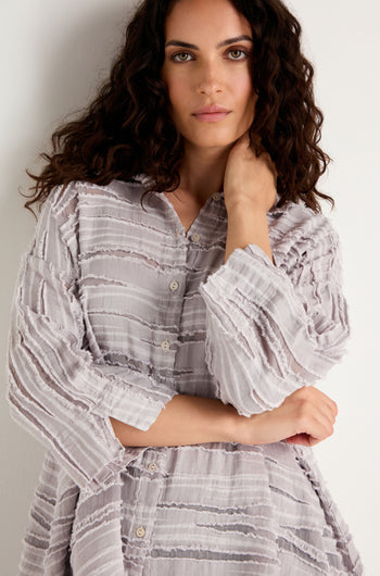 A woman with wavy dark hair stands against a white wall, looking at the camera with one hand touching her neck. She is wearing the Pearl Textured Stripe Flared Shirt, crafted from a luxurious linen-silk-cotton blend in a light color.