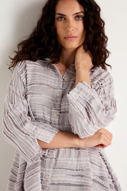A woman with wavy dark hair stands against a white wall, looking at the camera with one hand touching her neck. She is wearing the Pearl Textured Stripe Flared Shirt, crafted from a luxurious linen-silk-cotton blend in a light color.
