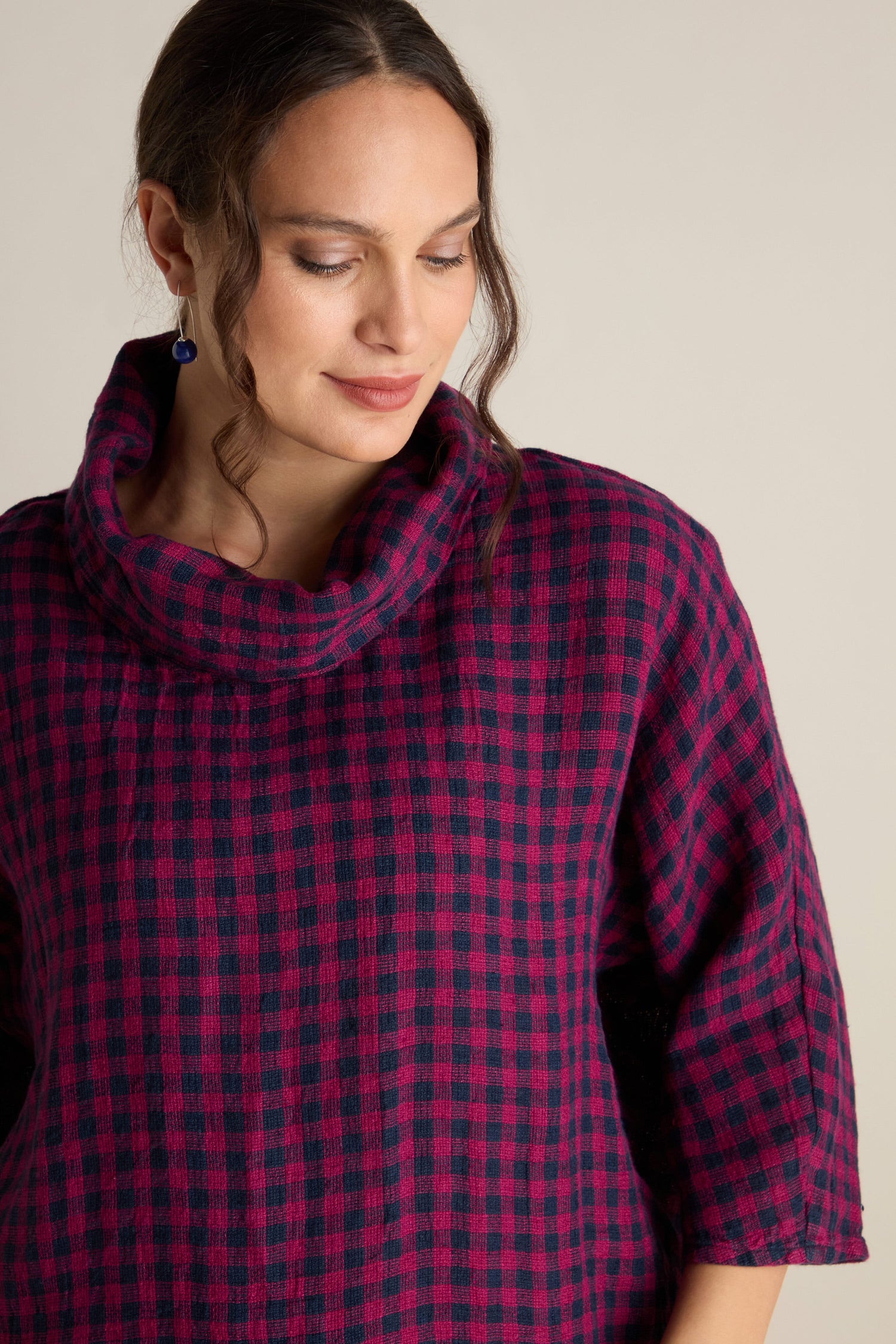 A woman with wavy hair in an updo wears the Linen Check Cowl Top, featuring a red and black checkered pattern and a boxy fit. She complements her look with blue earrings, looks down, and smiles slightly against a plain background, embodying the distinct Grizas style.