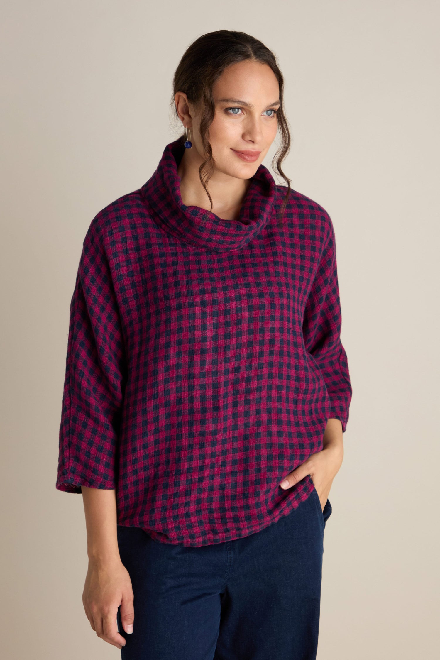 A woman wearing the Linen Check Cowl Top, which features a cozy, textured pink and blue checkered pattern, along with dark pants is standing against a plain background.