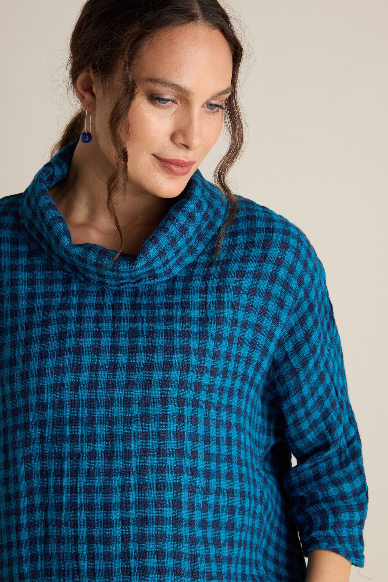 A person wearing the Linen Check Cowl Top, featuring a boxy fit with blue and black checkered pattern, reminiscent of Grizas style, has their hair partially tied back and hanging loosely on the sides while looking downwards with a neutral expression.