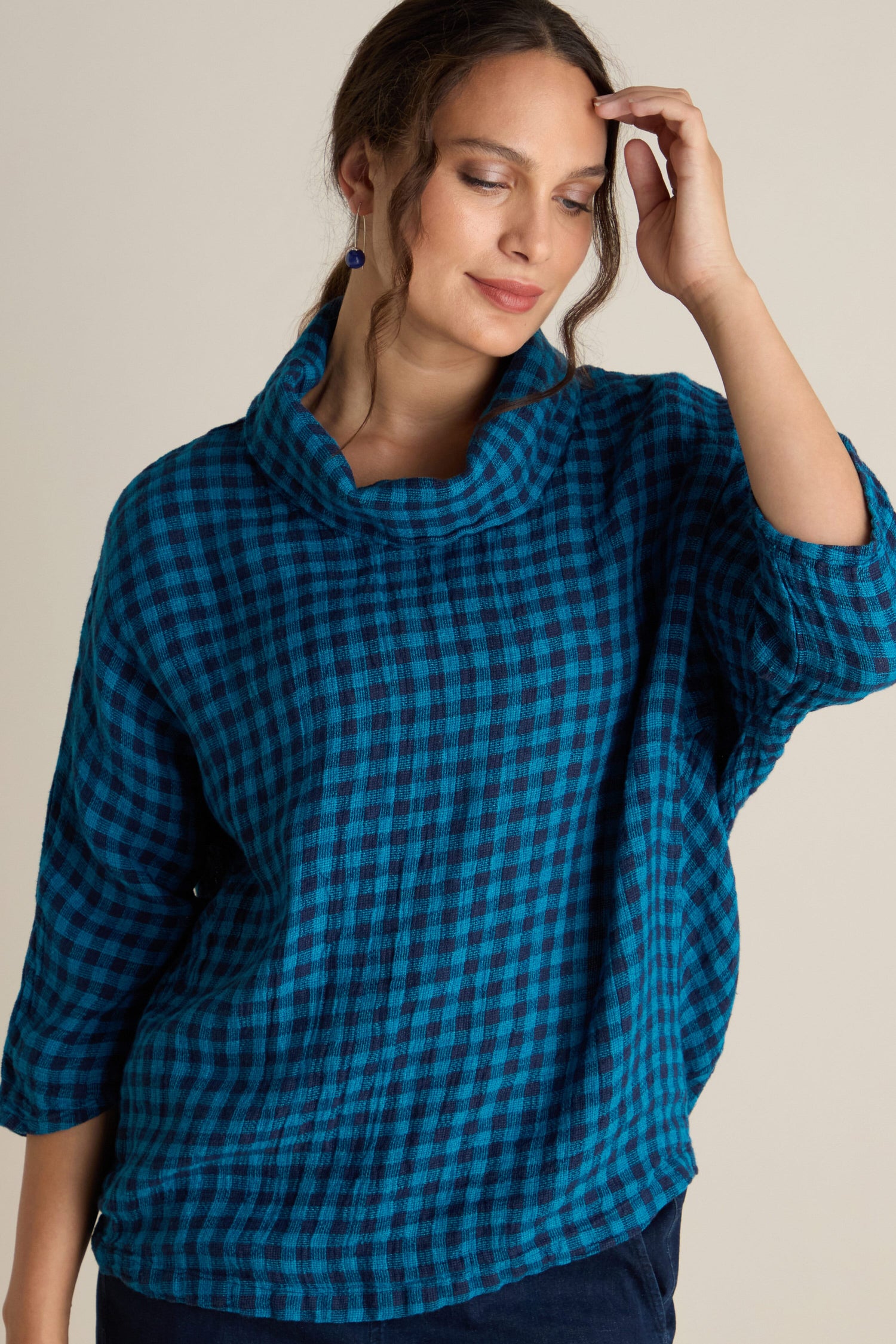 A person with long hair wearing a boxy fit Linen Check Cowl Top in blue and black poses with one hand touching their head.