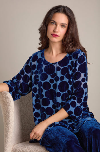 A woman seated in a beige chair wearing the Spot Devore Top, characterized by its blue velvet fabric adorned with large dark blue circles, looks at the camera.