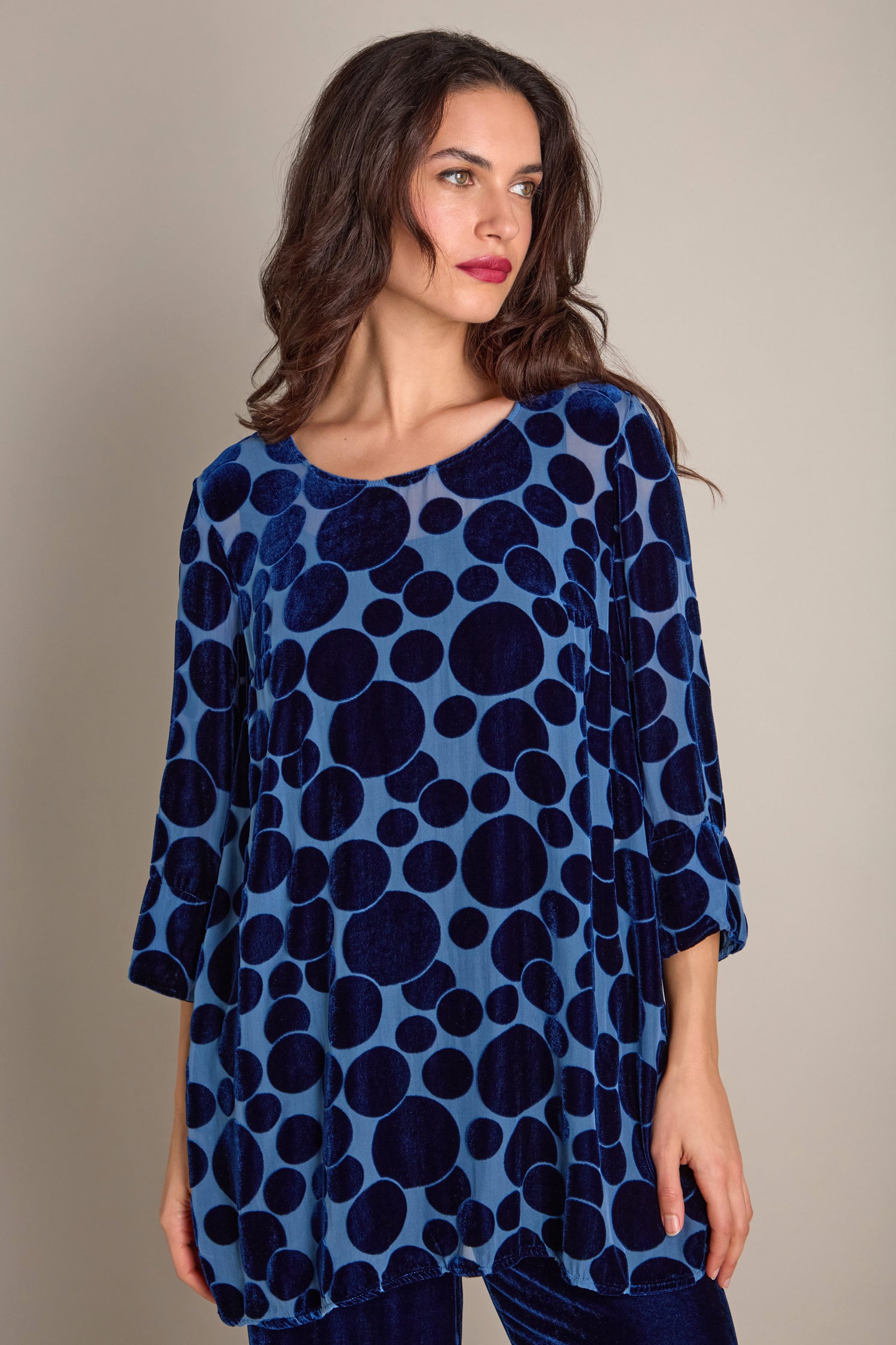 Against a neutral backdrop, a woman showcases the Spot Devore Top, adorned with large dark blue polka dots that effortlessly combine elegance and contemporary style.