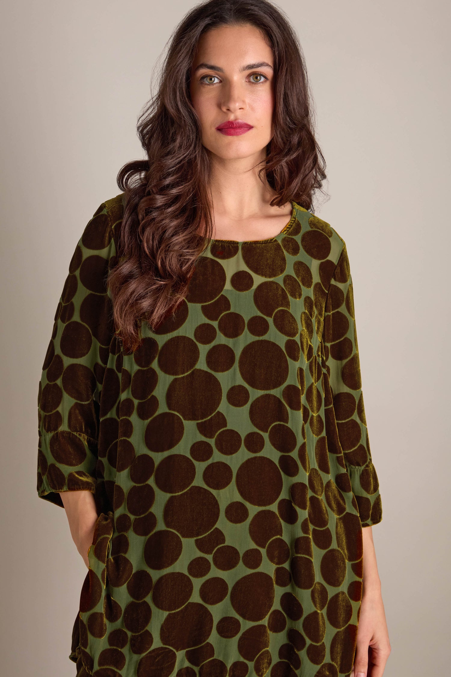 A woman with long hair wearing a Spot Devore Top in green velvet decorated with large dark brown polka dots stands elegantly against a plain background.