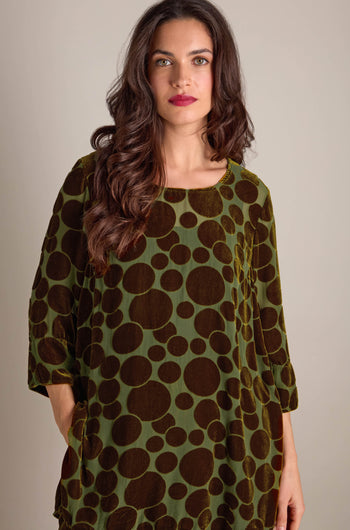 A woman with long hair wearing a Spot Devore Top in green velvet decorated with large dark brown polka dots stands elegantly against a plain background.