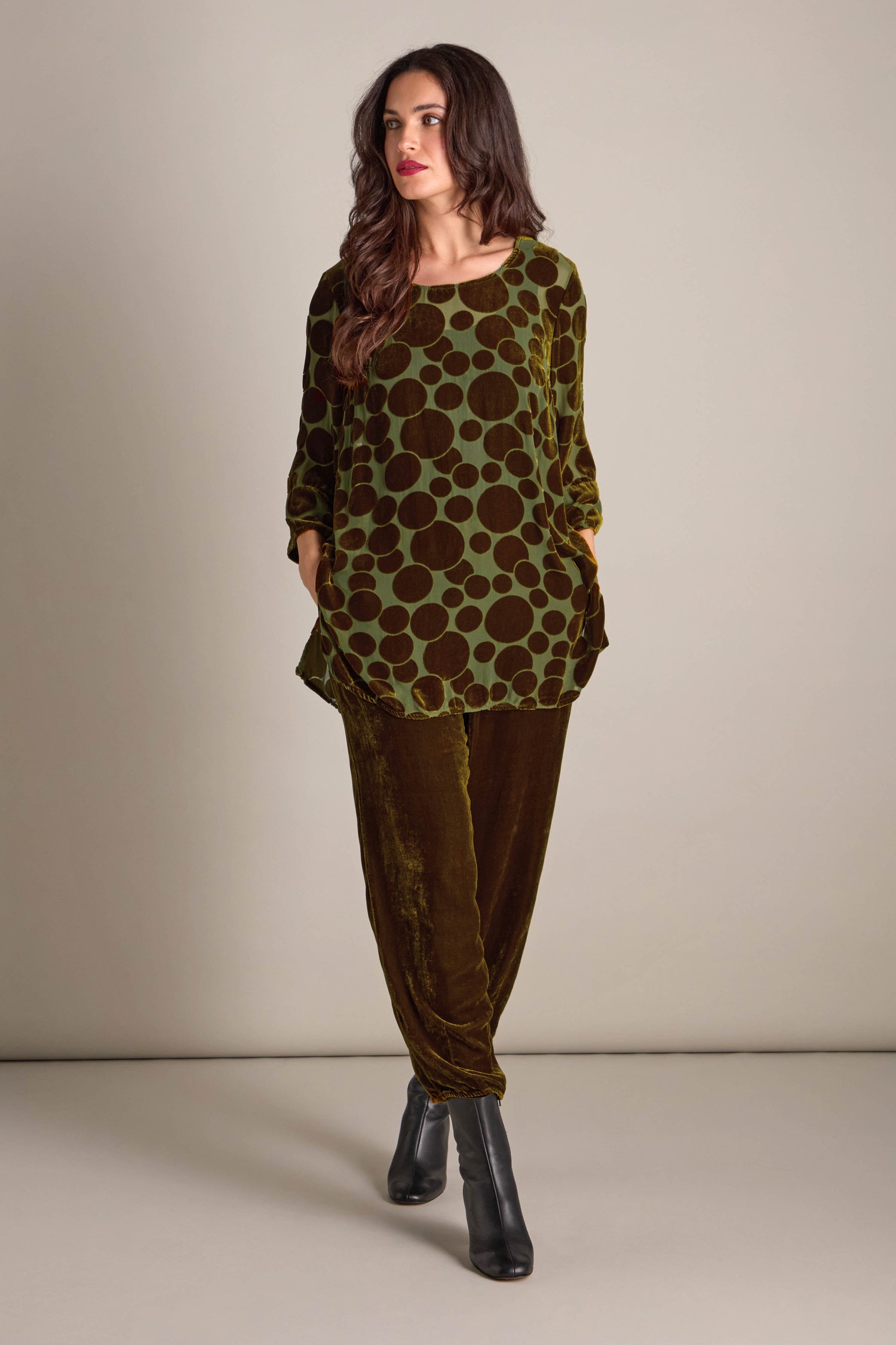 A person stands wearing the Spot Devore Top, a green velvet tunic with polka dots, paired with brown pants and black heeled boots, against a plain background.