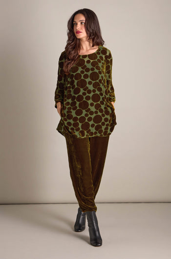 A person stands wearing the Spot Devore Top, a green velvet tunic with polka dots, paired with brown pants and black heeled boots, against a plain background.