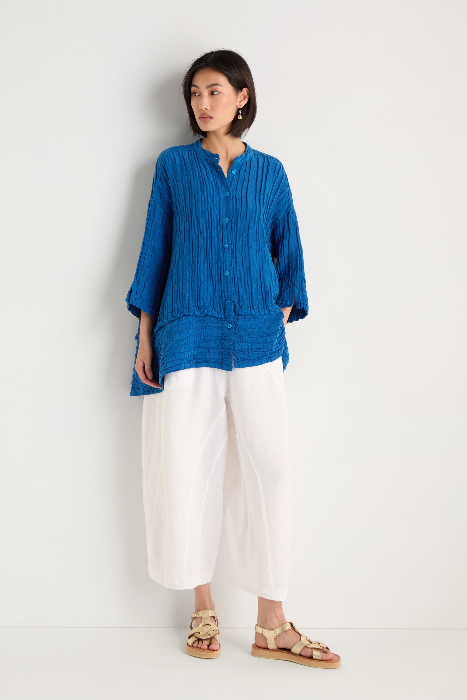 A woman stands against a white wall wearing the Panel Hem Silk Linen Shirt in blue with an oversized fit, paired with white wide-leg pants and beige sandals.