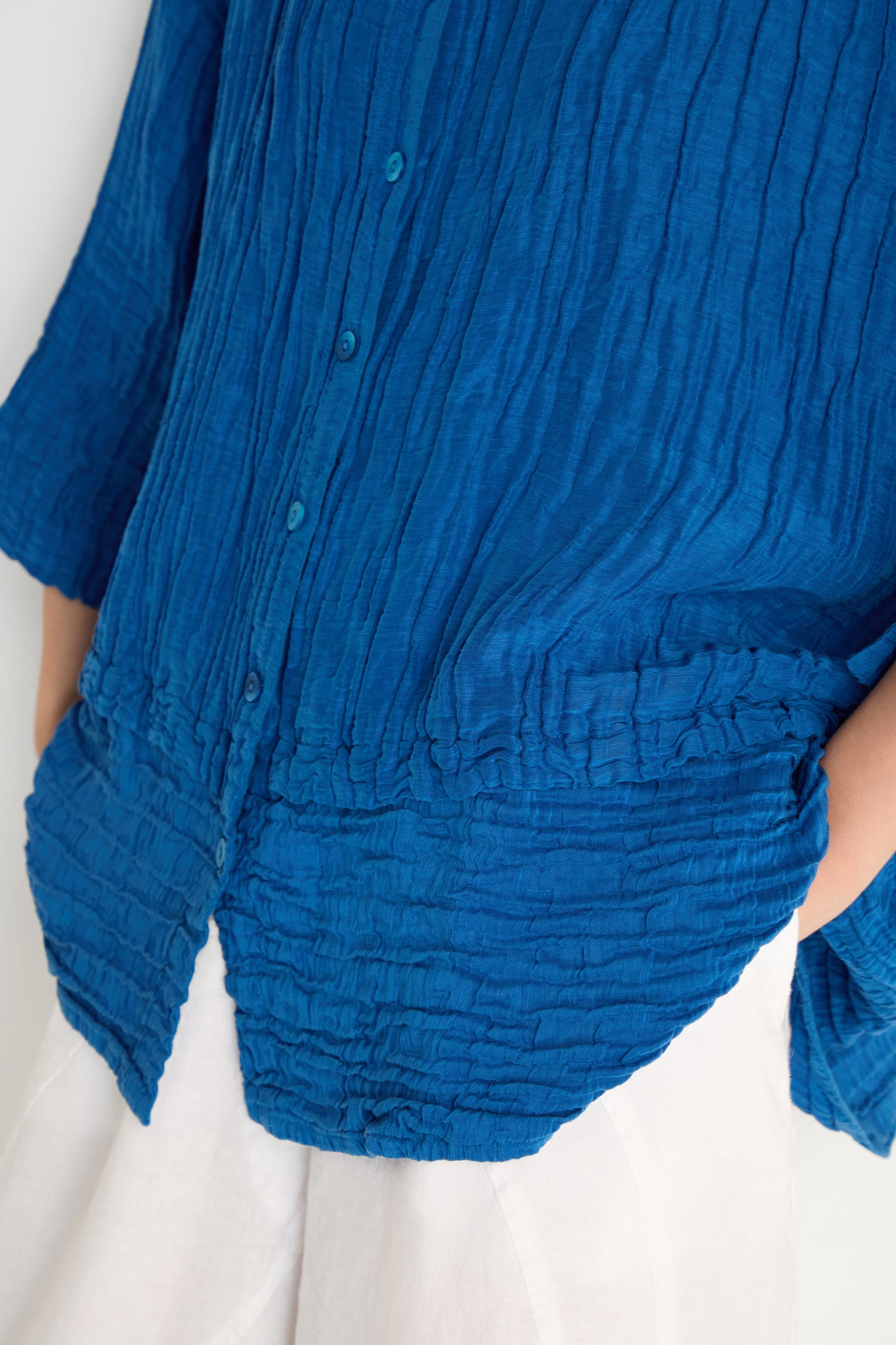 A person wearing an oversized Panel Hem Silk Linen Shirt in crinkled blue, with their hands in the pockets of white pants.