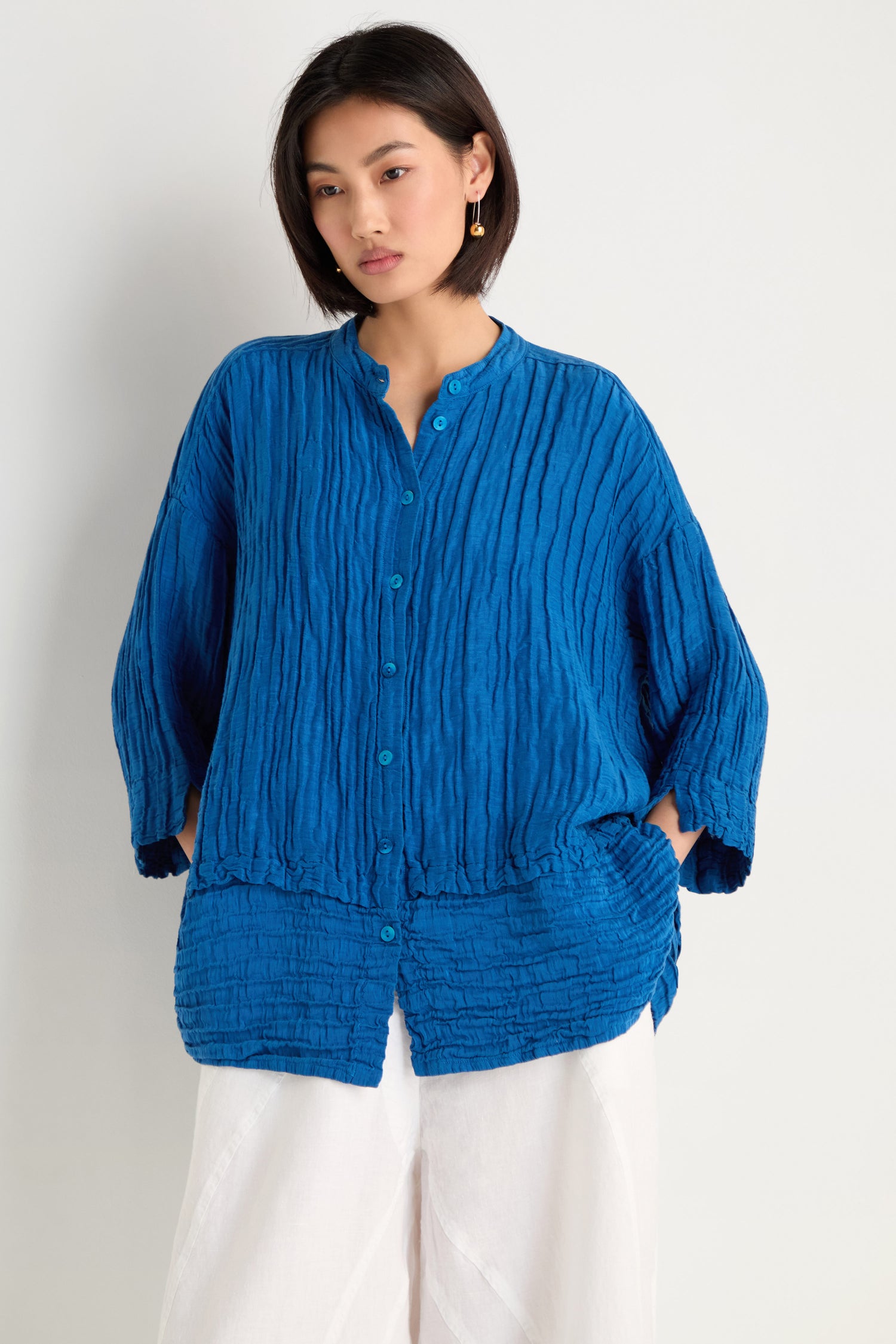 A woman stands against a plain background wearing the Panel Hem Silk Linen Shirt in blue, featuring breathable textured fabric, an oversized fit, and paired with white pants.