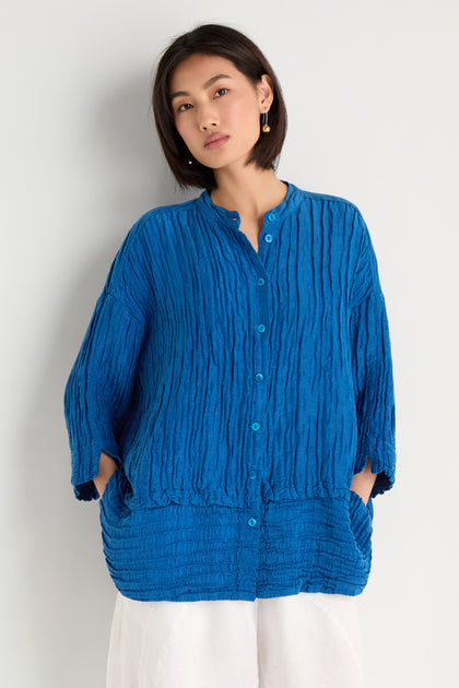 A woman wears the Panel Hem Silk Linen Shirt—an oversized, breathable blue top with long sleeves—paired with white pants and standing against a plain light background.