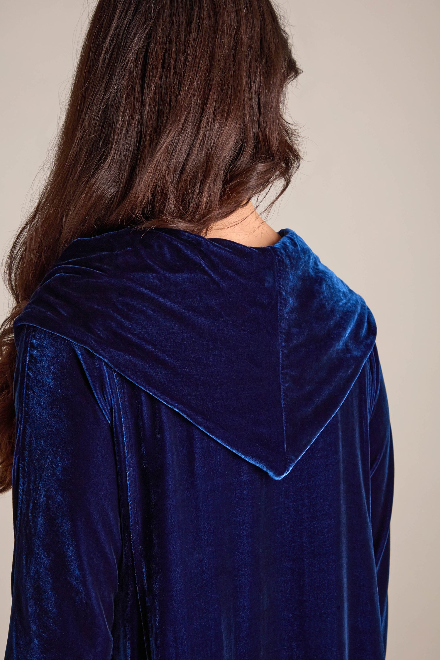 A person with long brown hair is elegantly modeling the Cowl Silk Velvet Dress, featuring a luxurious blue fabric and a hood, beautifully captured from the back against a plain background.