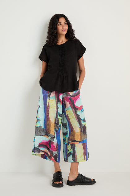 A woman stands against a plain wall wearing a Pleat Front Linen Top, colorful wide-leg pants, and black sandals, with her hands in her pockets.