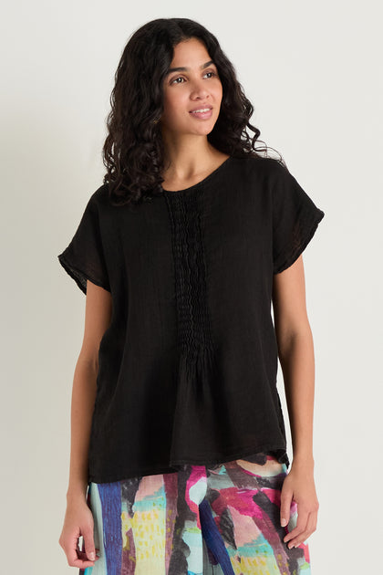 A woman with curly hair wears the Pleat Front Linen Top in black with a relaxed silhouette and front gathers, paired with colorful abstract print pants, standing against a plain light background.