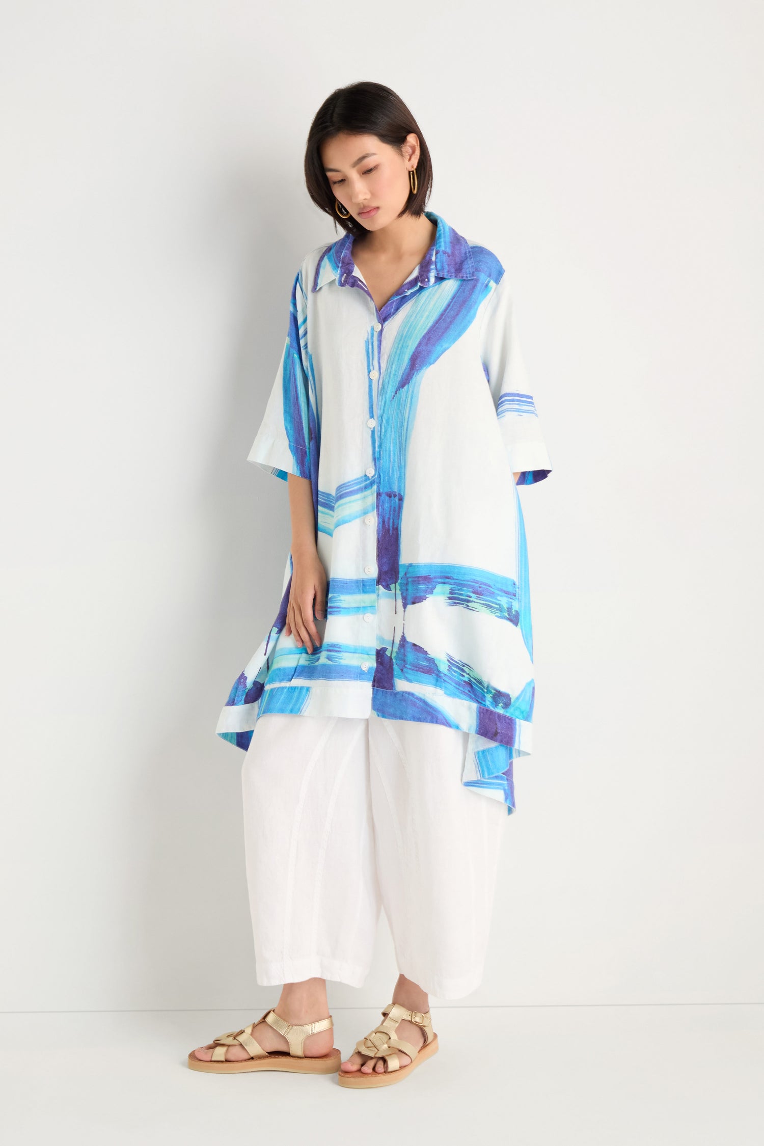 A woman models the Brushstroke Long Linen Shirt, featuring a loose blue and white pattern, styled over wide-leg white pants and beige sandals against a plain white background.