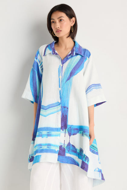A woman models the Brushstroke Long Linen Shirt, featuring a blue and purple brushstroke print, styled over white pants against a plain light backdrop.
