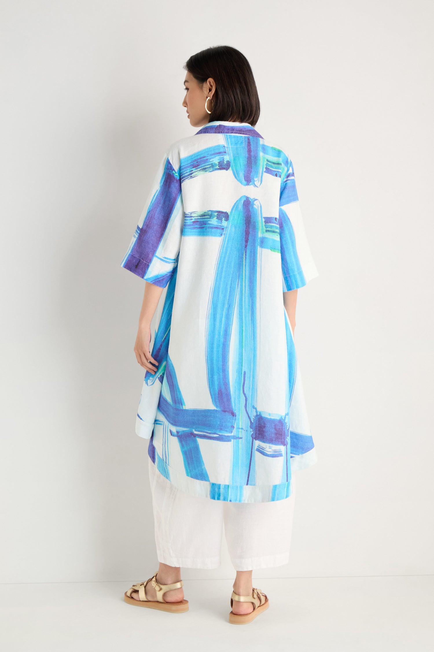 A woman stands against a white wall wearing the Brushstroke Long Linen Shirt with a bold blue print, paired with white pants and beige sandals, facing away from the camera.
