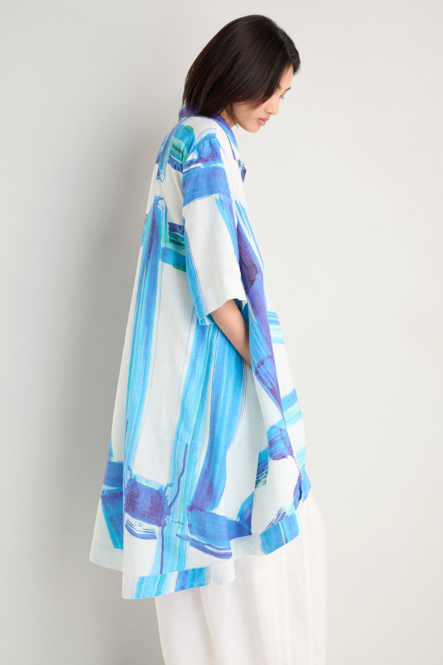 A person stands sideways against a plain light background, wearing the Brushstroke Long Linen Shirt—a loose, white longline shirt with blue brushstroke print—and white pants.