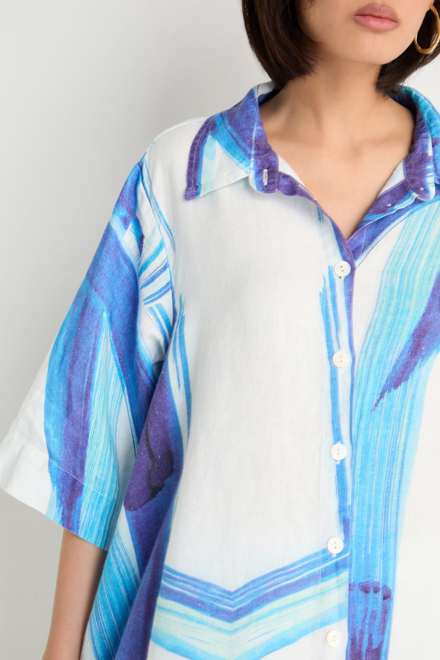 A woman models the Brushstroke Long Linen Shirt, featuring a loose fit and blue and purple brushstroke design; only her lower face and upper torso are visible.