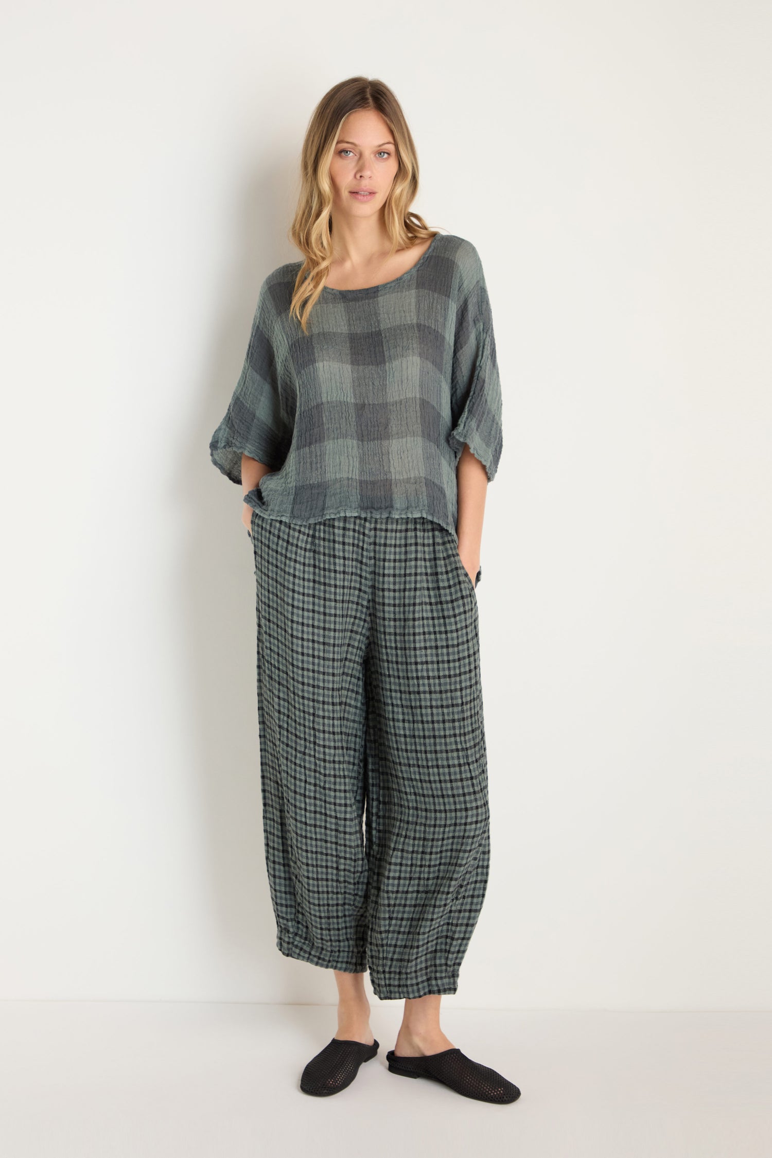 A woman stands against a white background wearing the Large Check Boxy Top in woven linen with wide pants in a matching large check pattern, paired with black slip-on shoes.