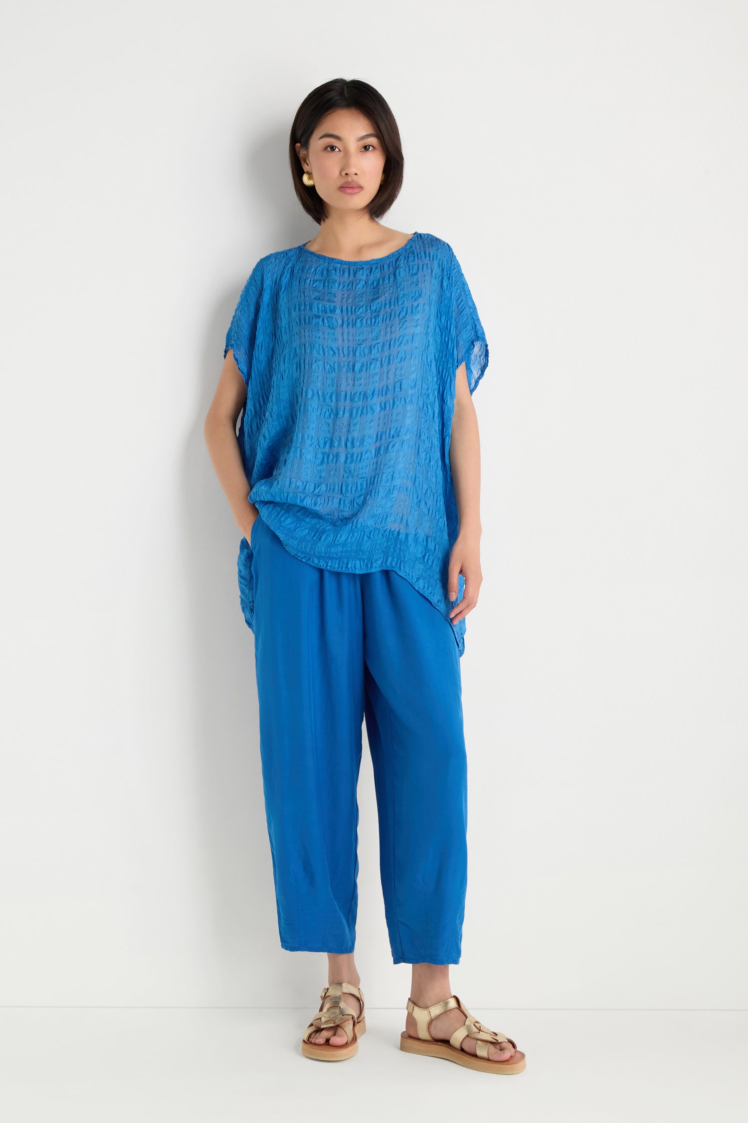 A woman stands against a white wall wearing the Sheer Crinkle Silk Boxy Top in blue, matching blue pants, beige sandals, and gold earrings.
