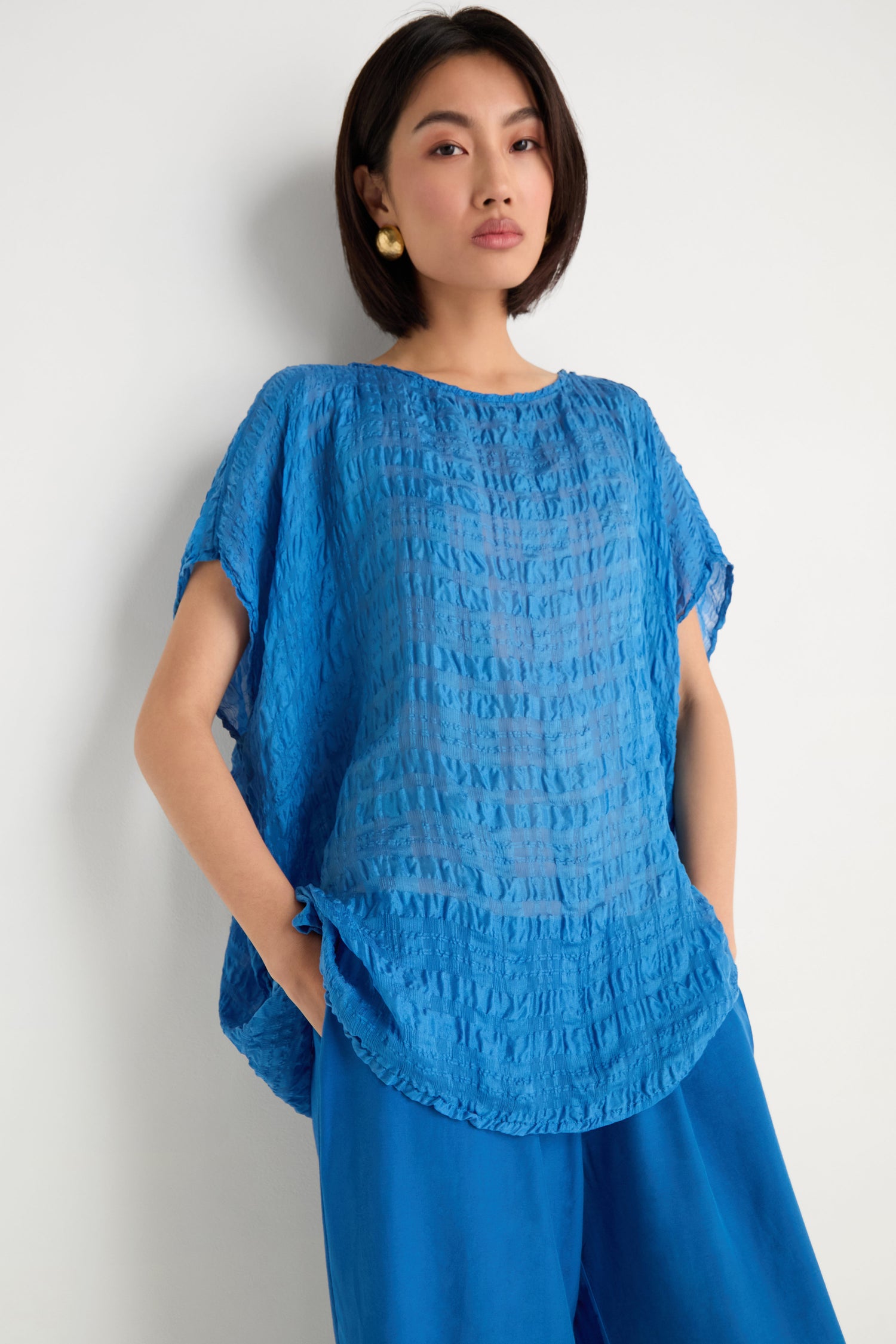 A person with short dark hair wears gold earrings and the Sheer Crinkle Silk Boxy Top, paired with loose blue pants, while standing against a white wall.